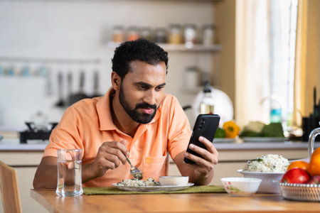 Indian middle aged man busy using mobile phone while eating lunch on dining table at home - concept of modern lifestyles, internet distraction and social media sharingの写真素材