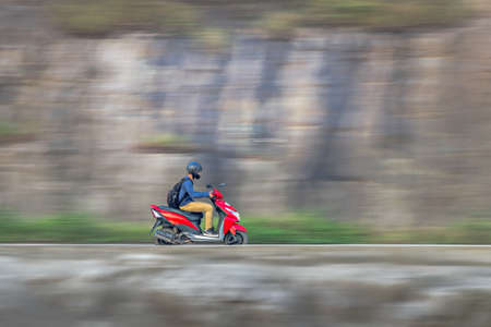 Motion blur image of a rider wearing helmet for safety, riding uphill on a red two wheeler moped.のeditorial素材