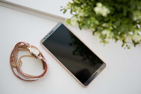 smartphone and women's jewelry lie on a white windowsill against a green flowerの写真素材