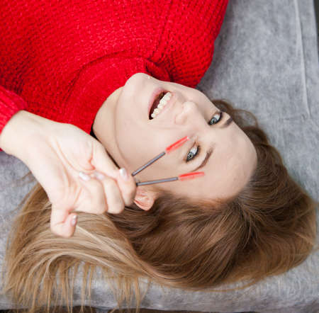 beautiful girl in a red sweater is lying, master eyelash extension with tweezers in her hands and brushes in her hairの写真素材