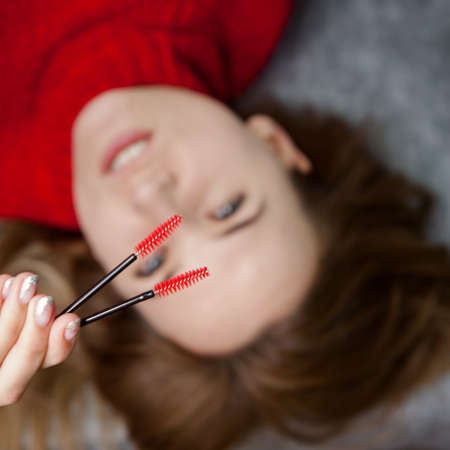 beautiful girl in a red sweater is lying, master eyelash extension with tweezers in her hands and...