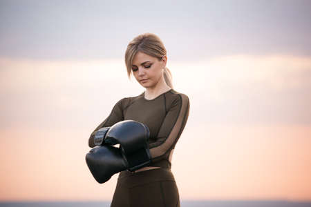 young adult athletic woman with long blond hair puts on black boxing gloves against the sunset skyの写真素材