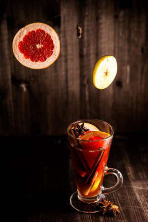 christmas mulled wine, in a transparent glass with fruit on a dark wooden background surrounded by flying spices and fruitsの写真素材