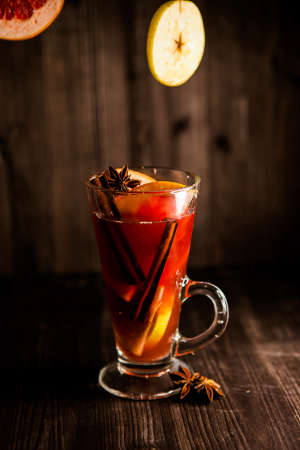 christmas mulled wine, in a transparent glass with fruit on a dark wooden background surrounded by flying spices and fruitsの写真素材