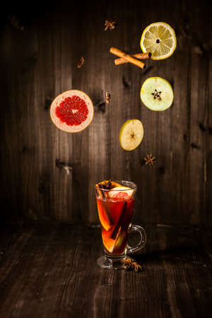 christmas mulled wine, in a transparent glass with fruit on a dark wooden background surrounded by flying spices and fruitsの写真素材