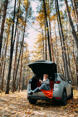 a blonde adult girl in a plaid shirt is drinking coffee sitting in the open trunk of a car. picnic in the car.の写真素材