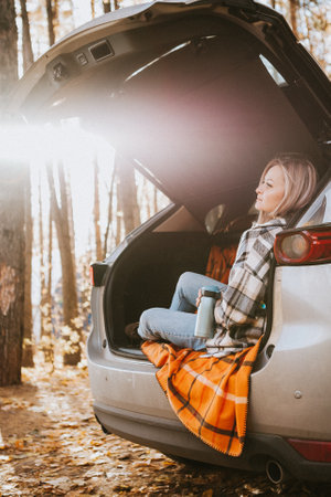 a blonde adult girl in a plaid shirt is drinking coffee sitting in the open trunk of a car. picnic in the car.の写真素材