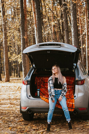 a blonde adult girl in a plaid shirt is drinking coffee sitting in the open trunk of a car. picnic in the car.の写真素材