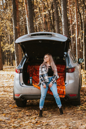 a blonde adult girl in a plaid shirt is drinking coffee sitting in the open trunk of a car. picnic in the car.の写真素材