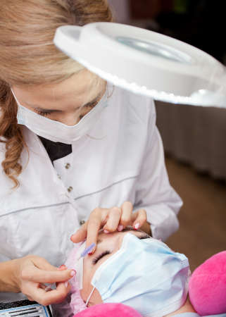 A girl makes the procedure for extending eyelashes during a pandemic, a master with tools in his...