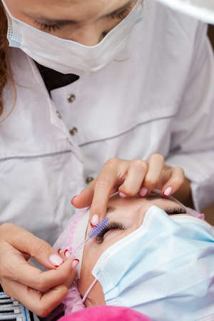 A girl makes the procedure for extending eyelashes during a pandemic, a master with tools in his...
