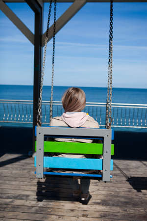 A girl with long blonde hair is sitting on a swing with a view of the sea, photo from the back....