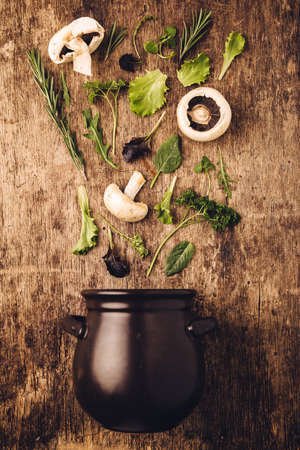 Layout of  fresh vegetables and mushrooms entering in the dark black pot against old wooden kitchen table background with nice, raw, texture. Nature spring time for healthy food. Creative concept of vegan food.の写真素材