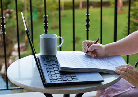 hand holding pen over paperwork with laptop on balcony represents work from home