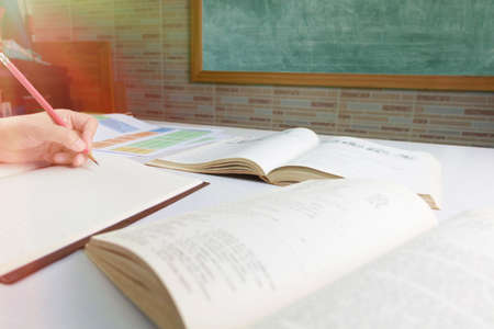 hand holding pencil over blurred textbook on table in front of blackboard in classroomの写真素材
