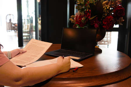 hand holding red pen over blurred paperwork on wooden table with laptopの写真素材