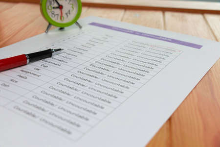 English grammar test sheet with red pen and clock on wooden table in classroomの写真素材