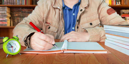 hand holding red pen about to write on notebook on wooden table with blurred bookshelf background