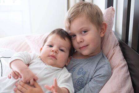 Toddler boy brother holding his newborn baby sister, sitting on the bed. Lifestyle
