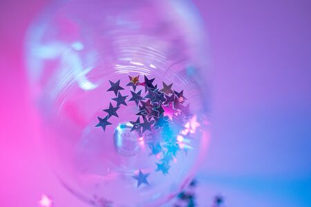 empty glass of wine with star shaped glitter confetti in pink and blue neon gradient lightの写真素材