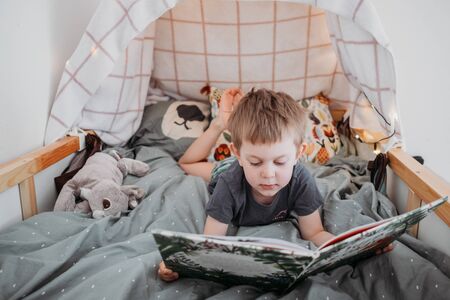 preschooler boy reading book laying in his bed. spending time at home. Lifestyle family conceptの写真素材