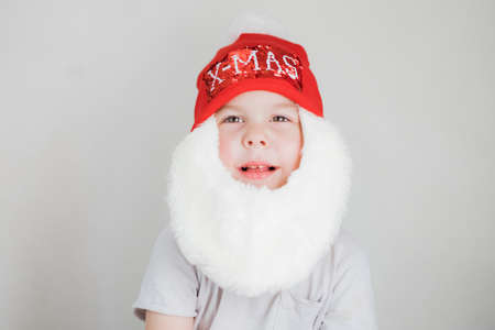 toddler boy in Santa Claus costume with beard and red hat on white background
