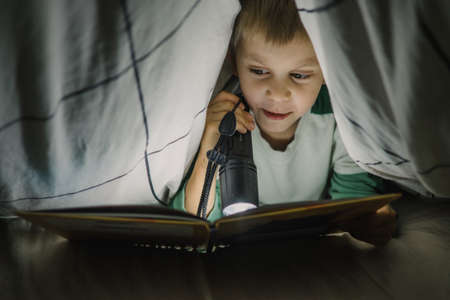 Little boy reading a book at night with lantern covered with blanketの写真素材