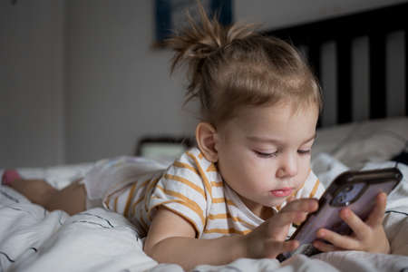 Little toddle girl playing with smartphone laying on the bed at home. Caucasian toddler girl.の写真素材
