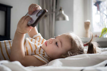 Little toddle girl playing with smartphone laying on the bed at home. Caucasian toddler girl.の写真素材