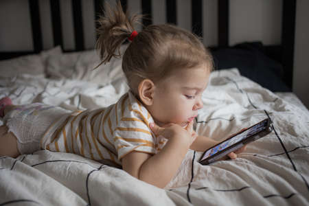 Little toddle girl playing with smartphone laying on the bed at home. Caucasian toddler girl.の写真素材