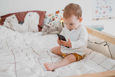 Infant girl sitting on bed and using smart phone in neutral tones nursery. Gender neutral baby nursery and clothesの写真素材