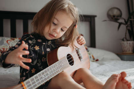 toddler girl playing ukulele guitar sitting on the bed at homeの写真素材