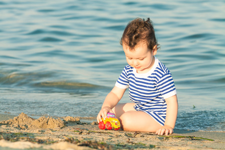 Adorable toddler with sailor shirt playing with a car at the edge of the water on a beach. Photo with untraditional color rendering for artistic lookの写真素材