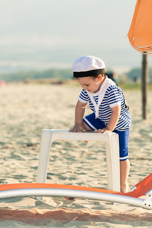 Playful toddler dressed as a sailor standing on a beach and climbing on a plastic table. Photo with untraditional color rendering for artistic lookの写真素材