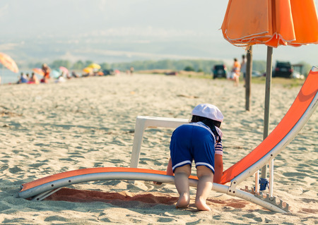 Sweet toddler dressed as a sailor climbing on a sunbed with view from his back. Photo with untraditional color rendering for artistic lookの写真素材