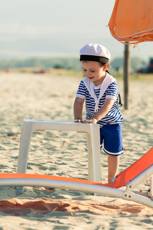 Sweet toddler dressed as a sailor standing on a beach near a plastic table and sunbed. Photo with untraditional color rendering for artistic lookの写真素材