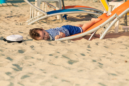 Toddler dressed as a sailor lying from a top of a sunbed on a beach and looking as he is drunk with his hat near him. Photo with untraditional color rendering for artistic lookの写真素材