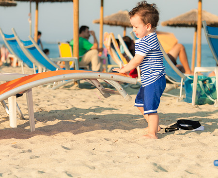 Toddler dressed as a sailor standing on a beach and playing with sunbed with other people in defocussed background. Photo with untraditional color rendering for artistic lookの写真素材