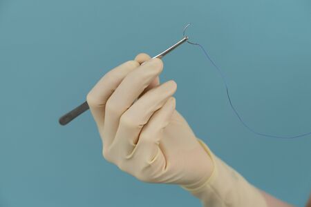 doctor woman holding a surgical thread with a needle in gloves with a mask on a blue backgroundの写真素材