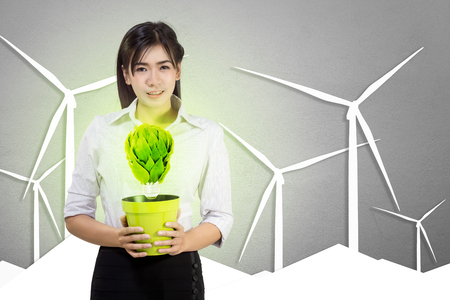 A woman holds a tree pot with an artificial light bulb made of leaves in wind turbine backgroundsの写真素材