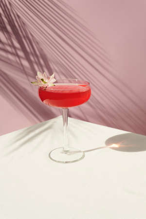 Pink cocktail decorated with a flower on the table with sun shadows. On a pink background with palm leaves, front view.の写真素材