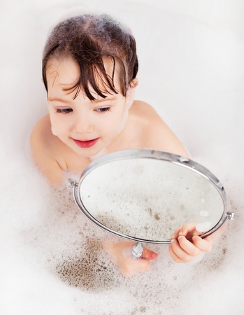 beautiful little girl taking a relaxing bath with foam and looking into the mirrorの写真素材