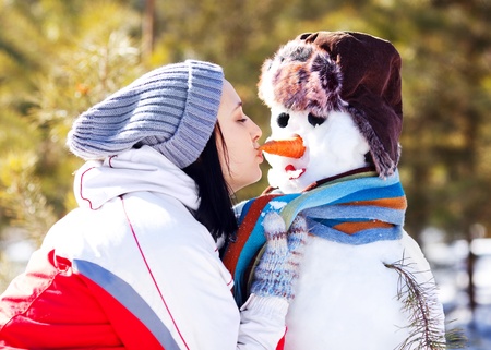 portrait of a pretty young brunette woman kissing a snowman outdoor on a warm winter dayの写真素材