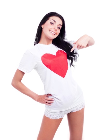 beautiful young woman wearing a T-shirt with a big red heart and pointing to the heart, isolated against white backgroundの写真素材