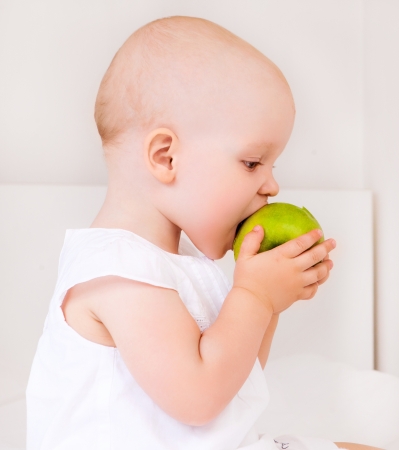 cute baby girl eating a green apple in bed at homeの写真素材