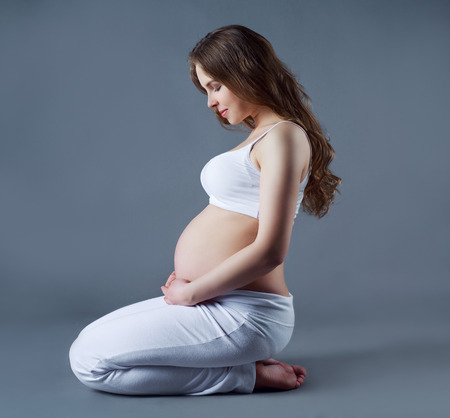 profile of a beautiful yougn pregnant woman sitting on the floor, against grey studio backgroundの写真素材