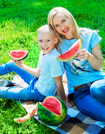 happy young mother and her ten year old son eatign watermelon in the summer parkの写真素材