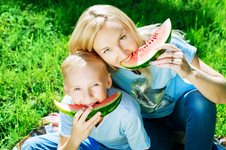happy young mother and her ten year old son eatign watermelon in the summer parkの写真素材