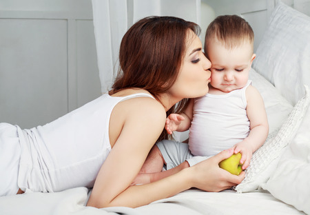 happy beautiful mother kissing her one year old son ,with a green apple, in bed at homeの写真素材
