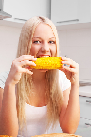 beautiful young blond woman eating corn at home in the kitchenの写真素材